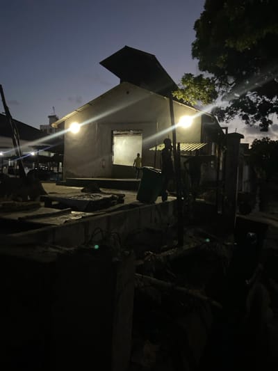 The image, captured at dusk or night, depicts an outdoor scene in Bagamoyo, Tanzania, dominated by a simple, light-colored building under artificial illumination. The sky is a deep twilight blue with visible clouds, suggesting early evening.
A prominent single-story building with a dark, corrugated metal roof stands on the left. Two bright outdoor light fixtures are attached to its facade, casting strong, ray-like beams across the scene and illuminating the building's exterior. Inside one of its window openings, which is brightly lit from within, a person wearing a light-colored (possibly yellow) shirt is standing, looking outwards or performing an activity.
To the right of the building, near one of the light sources, another figure is partially visible standing beside a green trash bin. The immediate surroundings of the building appear somewhat informal, with concrete walls, steps, and what seems to be debris or informal structures in the foreground and dark areas. Silhouettes of trees are visible against the sky in the upper portion of the image. The ground is uneven, characterized by various surfaces and possible rubble. No specific event beyond the presence of people in their daily environment is clearly discernable, and the atmosphere appears neutral. There is no visible text in the image.