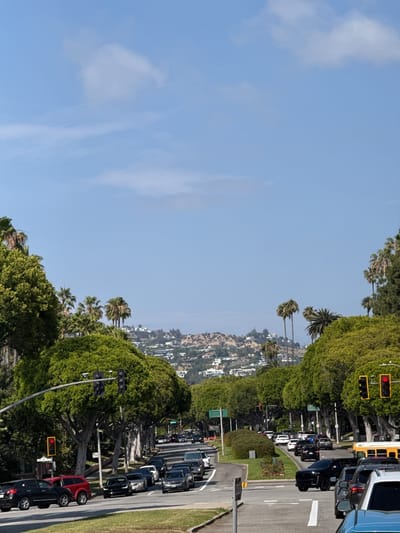 The image depicts a street scene in Beverly Hills, United States, characterized by a tree-lined avenue with moderate vehicular traffic. The road stretches towards distant hills dotted with residential structures. Traffic lights are visible along the avenue. The sky is light blue with sparse clouds. A median strip divides the road in the foreground.