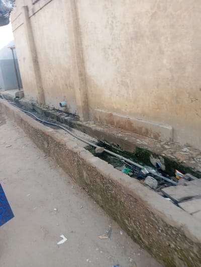 An exterior shot captures a scene of a dirty drainage ditch running alongside a textured, light-brown wall. The ditch is filled with murky water, debris, and green algae. A gray pipe extends from the wall into the ditch, and another hose-like pipe runs along the length of the ditch, partially submerged. The ground beside the ditch is made of compacted dirt and scattered litter. A small portion of a blue fabric with a red swirl pattern is visible in the lower-left corner of the frame, suggesting a person is present but out of view. The lighting suggests it is daytime. The location is identified as Gwagwa, Nigeria.