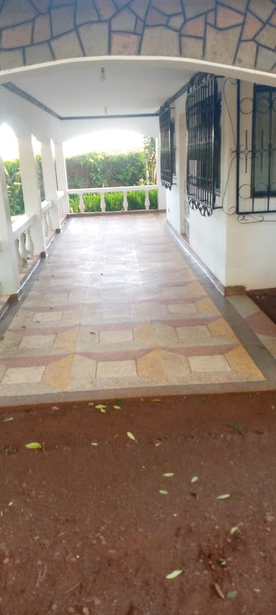 A covered porch extends along the side of a house. The porch floor has tan, cream, and mauve geometric tiles. White pillars support the roof. Wrought iron covers the windows. Beyond the porch, a clipped green hedge is visible. The ground below the porch is bare soil with scattered small green leaves. The overhead roof of the porch is white.