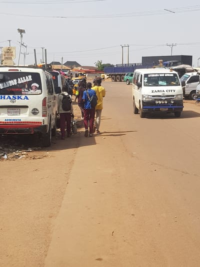 The image captures a bustling street scene in Kufena, Nigeria, on a clear, sunny day. The perspective is from the roadside, looking down a street filled with vehicles and pedestrians. The road surface appears to be a mix of asphalt and reddish-brown dirt.
In the foreground, on the right side of the road, several people are walking. One person in a yellow top and dark pants walks away from the viewer, while another in a blue shirt and dark pants follows. Shadows stretching from the people and vehicles suggest it is daytime, possibly morning or afternoon.
The road is lined with various vehicles. A prominent white commercial van, possibly a Toyota Hiace, is seen from the rear on the left side of the frame. Its rear window features text including "MANZON ALLAH" and a "TOYOTA" logo, along with "HASKA" in blue letters on its tailgate. A license plate "598XS" is visible, and the bumper has "ADMIMUTA" printed on it. Further down the street, another white truck or van is visible, displaying "AYIMANA DAN AHMADI" and "ZARIA CITY" on its front windshield. Its front bumper has a blue guard, and a license plate "301 678JA" can be faintly seen. This vehicle also has "PISTON'S" written on it.
Beyond these vehicles, a line of other cars, vans, and large trucks with blue trailers extend into the distance, indicating traffic or a busy parking area. Power lines and utility poles are visible against the bright sky, stretching along the side of the road. On the left side of the road, a street sign for "NASHUKA VENTURES LIMITED" can be identified. The general atmosphere suggests a lively urban or semi-urban environment.