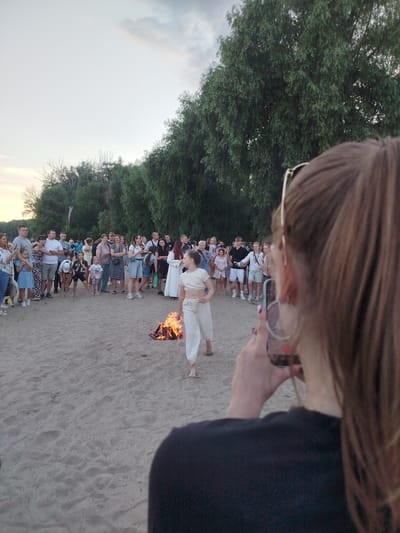 An outdoor gathering on a sandy beach in Chernihiv, Ukraine, features a bonfire with flames in the foreground. A young girl in white attire is captured in motion near the fire, her back to the viewer. A crowd of people, including adults and children, are gathered around, watching the event. A woman in the immediate foreground is taking a photo of the scene with her phone. The background is dominated by lush green trees and a pale sky, suggesting it is late afternoon or early evening. The overall atmosphere appears festive and communal.
