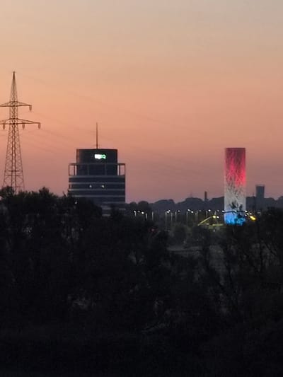 The image depicts a nighttime cityscape with a dramatic sky. The sky is a gradient of soft orange and pink hues, suggesting either a sunset or sunrise. Dominating the skyline are several structures, including a tall, slender obelisk-like building illuminated with red and white lights, possibly representing a national flag. Adjacent to it, other buildings are visible, including a multi-story office building with lit windows and a prominent green logo that appears to be a stylized letter "G" or "O."
In the foreground, a large, dark silhouette of trees obscures part of the scene, adding depth and contrast to the illuminated structures. Further to the left, a tall power pylon with visible wires stands against the twilight sky, indicating the presence of electrical infrastructure. The overall scene suggests an urban or industrial area at dusk or dawn. The lighting on the obelisk is particularly striking, creating a festive or patriotic atmosphere. The mood is serene yet dynamic, with the interplay of light and shadow. No people are visible in the image.