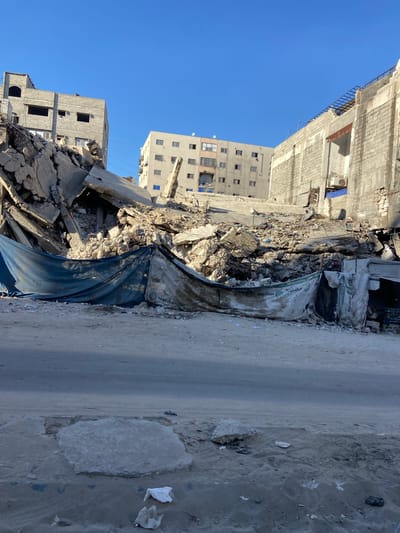 This is a vertical, eye-level, medium shot of a street in Gaza, Palestinian Territories, where buildings have been severely damaged or destroyed. The scene depicts widespread destruction with large piles of rubble, concrete debris, and twisted metal filling the foreground and midground. Tarps, one dark blue and another white with green lettering, are draped over some of the debris, possibly for temporary covering or as part of a salvage effort. A paved road runs along the right side of the image, partially obscured by debris and sand.

In the background, several buildings stand, some appearing intact with windows and balconies, while others show visible damage. One building on the left appears to be under construction or renovation, with exposed rebar and unfinished concrete. The sky is a clear, bright blue, suggesting a sunny day, likely mid-morning or afternoon. The overall mood conveyed is one of devastation and the aftermath of conflict.

Visible text on the white tarp reads "segezha group" in green lettering.