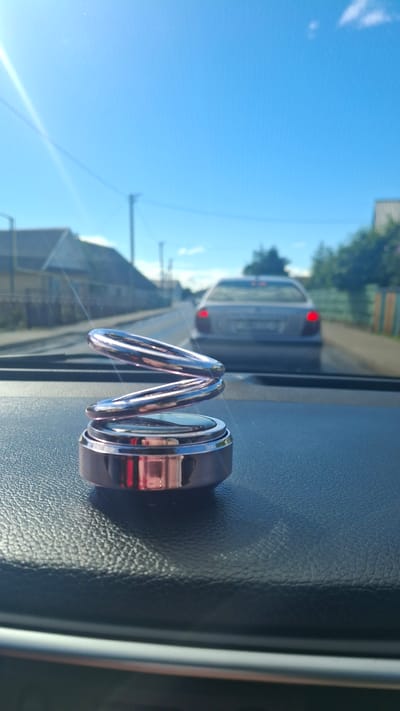 The image, captured from the interior of a vehicle, shows a shiny, coiled air freshener placed on the dashboard. The air freshener is rose gold in color and has a spiral design. The vehicle is in motion on a paved road, indicated by the blurred scenery in the background. Ahead, another car with its brake lights illuminated is visible. The sky is a clear blue with a bright sun casting a lens flare across the image. Buildings and trees line the roadside. This scene is likely set in Stowbtsy, Belarus, given the architecture and vegetation typical of the region. The overall impression is one of a bright, sunny day in a suburban or rural area.