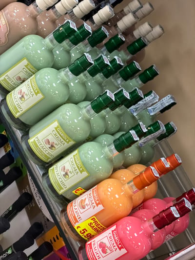 This is a medium shot, overhead, angled photograph of a shelf filled with bottles of liqueur. The bottles are arranged in rows, with some in front of others, and they are tilted. The liqueur bottles are of various colors, including pale green, pink, and orange. Most of the bottles have labels that read "GIAROLA CREMA" followed by the flavor, such as "PISTACCHIO," "MELONE," or "FRAGOLA." Some of the bottles also have prices displayed on them, such as "€ 5,99." The background is a neutral, light brown color. The overall impression is of a retail display of alcoholic beverages. There are no people visible in the image. The setting appears to be a store or a liquor shop. The lighting is bright and even. There are some other objects in the background, but they are out of focus.