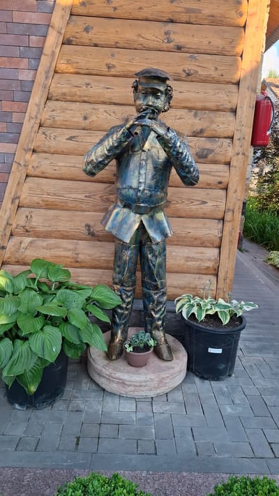 This is a medium shot of a metal sculpture of a man, presented in a vertical orientation. The sculpture, made of dark metal with iridescent blue and green highlights, depicts a man in a military-style uniform with a cap. He is posed with his hands at his sides, seemingly walking or standing at attention.

The sculpture is placed on a round, beige, stone-like base, which itself rests on a dark grey brick pathway. To the left of the sculpture is a rustic wooden wall, constructed from vertical planks. At the top of the wooden wall, a red object, possibly a fuel canister or a toolbox, is visible.

In the background, to the right of the sculpture, a large black planter holds a variegated hosta plant. Another smaller, brown pot with a small succulent is placed on the stone base, between the sculpture's feet. In the foreground, to the right and bottom of the frame, more lush green hosta plants are visible, along with a hint of a dark plastic pot. The scene is set outdoors, with a mix of paving stones, wooden structures, and greenery suggesting a garden or an outdoor decorative area.

The lighting indicates it is daytime, likely sunny, casting subtle shadows. The overall mood is one of outdoor display and decoration. There is no discernible text in the image, apart from a small white label on the large black planter, which is too blurry to read. The location context provided is Gyulagarak, Armenia.