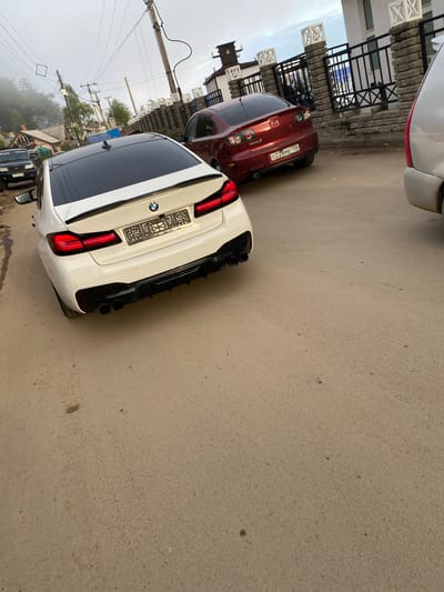 A white BMW sedan is parked on a narrow asphalt road in Listvyanka, Russia, its rear end facing the camera. The car has a black spoiler and black quad exhaust pipes. To the left of the BMW, a dark-colored SUV is visible. Further down the road, a red Mazda sedan is parked, displaying a Russian license plate that reads "O226ME 138". Behind the cars, a stone wall with decorative pillars and black metal fencing separates the road from a building. Utility poles with numerous wires are situated on the left side of the road, and a foggy, treed hillside is visible in the background. The sky is overcast.