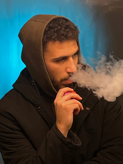 A man exhales a plume of white vapor from a pink device held in his hand. He wears a dark hooded jacket. The hood is pulled up. His face is partially visible, showing curly hair, a beard, and closed eyes. The background features an indistinct blue color gradient. The image is oriented at an angle.