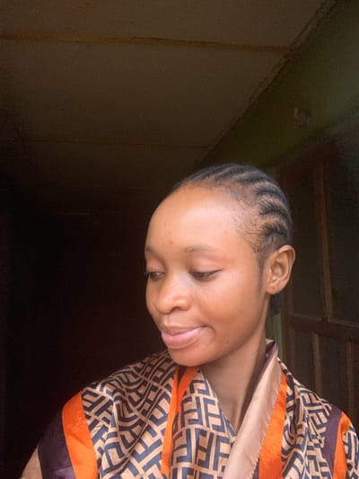 The media file features a close-up, portrait-style shot of a young Black woman with her eyes closed, appearing to be resting or in a moment of reflection. She has her hair styled in neat cornrows that frame her forehead. Her head is tilted downwards and to her right, with her face partially obscured by a patterned fabric draped around her. The fabric is a blend of warm beige and brown with an intricate geometric design in dark blue, accented by bright orange stripes along the edge.
The lighting suggests an indoor setting, possibly with natural light coming from a window or an opening to the side, casting shadows that create a sense of depth and intimacy. The background is dark and appears to be a wall, with some texture visible, possibly due to an aged or rustic finish. There are also hints of wooden structures above, suggesting a ceiling or overhead beams.
The woman's expression is serene, with a slight smile playing on her lips, conveying a feeling of peace or contentment. The overall mood of the image is calm and personal. There is no visible text in the media file. The location context of Shagamu, Nigeria, might imply a residential setting within the region. The time of day is not explicitly indicated, but the lighting does not suggest harsh midday sun, leaning more towards softer, ambient light.