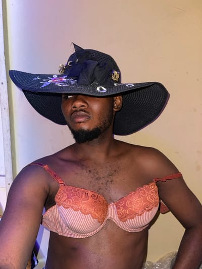 An adult individual, appearing male, with dark skin and a trimmed beard, is depicted indoors in Samaru, Nigeria. The subject wears a wide-brimmed black straw hat decorated with a black fabric bow, gold-colored embellishments, and iridescent accents, which partially obscures the upper face. The subject also wears an orange-peach colored bra with lace trim and subtle vertical stripes; the right shoulder strap is slipped down the arm. The subject's chest exhibits visible body hair. The pose is a slight turn to the right with a direct gaze. The background consists of a plain, light-colored wall with a visible light gradient from bluish-purple on the left to yellowish-beige on the right. A crumpled white material is partially visible in the bottom right corner.