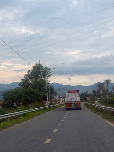 An eye-level, medium shot captures a red truck driving away from the camera on a paved road in Vietnam. The truck is carrying large, light-colored rectangular objects, possibly stone slabs. The truck's brake lights are illuminated, indicating it is either slowing down or stopped. The road has yellow dashed lines marking the lanes. On the left side of the road, a metal guardrail separates the road from a grassy embankment and vegetation. In the background, a large, leafy tree stands prominently, and beyond that, rolling hills covered in vegetation and mist are visible under a cloudy sky. Power lines crisscross the sky. On the right side of the road, there are more trees, including palm trees, and some buildings. Traffic signs are also visible along the road. The overall scene suggests a rural or semi-rural setting in Vietnam, likely during overcast weather, perhaps at dawn or dusk given the lighting.