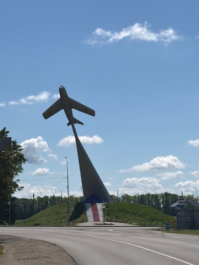 This media file shows a monument of a Soviet-era jet fighter plane, likely a MiG-17, displayed against a bright blue sky with scattered white clouds. The plane is mounted on a tall, pointed support structure, appearing to ascend. Red stars are visible on the wings of the plane. The scene is oriented vertically in the image.
In the background, to the right of the plane and support, there is a road with a white dividing line curving away. Along the roadside, there is a grassy slope covered in dense green foliage and trees. Further in the background, a small bus stop shelter with a blue sign featuring a white bus icon is visible. A building with red and white striped panels is situated behind the bus stop. The overall setting suggests an outdoor public space, possibly a roadside monument or park area in Leningradskaya, Russia. The time of day appears to be daytime, and the weather is clear with a few fluffy clouds. No people are visible in the frame.