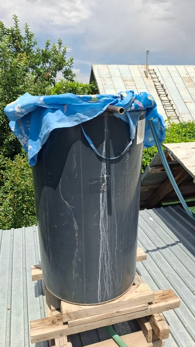 A large, dark gray plastic barrel sits on a makeshift wooden platform on a corrugated metal roof. The barrel is partially covered with a blue cloth adorned with white patterns. A metal pipe protrudes from the top of the barrel, and a blue hose is visible running into the barrel from the right. Water stains streak down the side of the barrel. In the background, trees, a house with a tin roof, and a ladder are visible under a cloudy sky. The scene is likely in the city of Shubarssu, Kazakhstan.