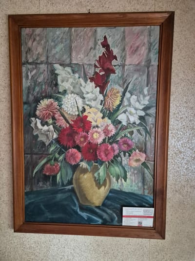 A framed oil painting depicts a bouquet of flowers in a golden vase on a dark green tablecloth. The background is a grey, tiled wall. A white and red business card is placed on the lower right corner of the painting. The painting is hanging on a beige, textured wall. The business card contains information about "Pflegeberatung Birgithe Scholz" and contact details for Würzburg, Germany. The location is identified as Rottendorf, Germany, based on the business card.