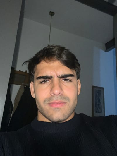 The media file features a close-up, portrait-style image of a young man, likely a selfie, captured indoors in Avellaneda, Argentina. The man's head is tilted, and he gazes directly at the viewer with an intense, somewhat serious or contemplative expression.
He has dark brown hair, dark brown eyes with prominent, well-groomed eyebrows, and visible stubble or a short beard on his chin, jawline, and upper lip. His lips are naturally full and possess a reddish-pink color. He is dressed in a dark, possibly black, ribbed or textured sweater or top that covers his neck and shoulders.
The background indicates an indoor setting, likely a home, with soft, dim lighting suggesting evening or night. A light-colored wall (off-white or light grey) occupies most of the background. On this wall, a dark-framed picture or mirror is discernible in the upper right. To the upper left, a small, dark circular object mounted on a thin rod, possibly a light fixture or ceiling element, is visible. The overall ambiance is quiet and subdued. No visible text is present in the image.