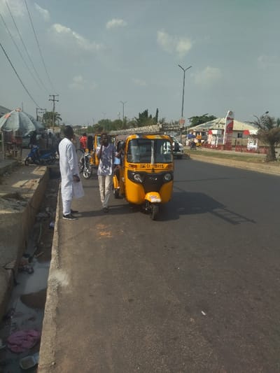 The scene is a street in New Karu, Nigeria. Three men are standing on the sidewalk next to a yellow auto rickshaw. The rickshaw is parked on the street, and the men appear to be interacting with the driver. The driver is talking on a cell phone. Behind the men, there is a drain on the side of the road. The men are wearing casual clothing. There are some other vehicles visible in the background, including a white car and a motorcycle. There are power lines overhead. The road is paved and appears to be in good condition. The sky is cloudy. The image was taken from a low angle and the scene is in focus. The lighting is sunny and bright. A building can be seen in the distance, behind the yellow rickshaw. The building has a red and white sign. There are trees and vegetation on the opposite side of the street. The scene is typical of a busy street in New Karu.
