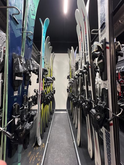 The image depicts a ski shop in Val-d'Isère, France, located at the coordinates 45.442, 6.977. Two rows of skis are displayed on vertical metal racks, aligned perpendicular to the floor. The skis are organized according to size and type, with black, white, yellow, and blue skis all present. Several pairs of ski bindings are attached to the skis, as are black rubber straps. The shop is illuminated by fluorescent lights, and the floor is a black and gray patterned tile. The scene is empty of people.