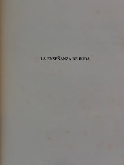The image is a faded book cover or page with the text "LA ENSEÑANZA DE BUDA" printed vertically in black. The background is a light, off-white color with faint yellowed spots. The paper appears old and slightly worn around the edges. The text is centered on the left side of the image.