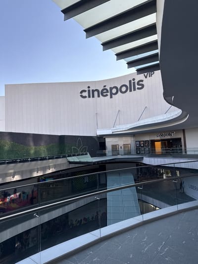 The image depicts a multi-level modern commercial complex, specifically the Cinépolis VIP cinema at Plaza Mayor in León, Guanajuato, Mexico. The perspective is from an upper level, looking down and across at lower levels and the cinema facade.
In the upper portion, a clear blue sky is visible. A large, curved, light-colored building wall dominates the background, featuring the dark grey lettering "cinépolis VIP." Above and to the right, a multi-paneled overhead structure with dark crossbeams and transparent elements is partially visible, indicative of a modern roof or canopy.
Below the main "cinépolis VIP" sign, a smaller entrance also marked "cinépolis" displays movie posters behind glass. To the left of this entrance, a textured wall features a vertical garden composed of dark and lighter green artificial foliage, including a stylized leaf pattern.
The foreground and mid-ground are characterized by multiple architectural levels with dark, patterned flooring and contemporary glass railings with stainless steel handrails. Reflections of structural elements and distant commercial spaces are visible on the glass and polished floor surfaces. No individuals are clearly identifiable within the visible area of the complex, which appears well-lit by natural daylight.