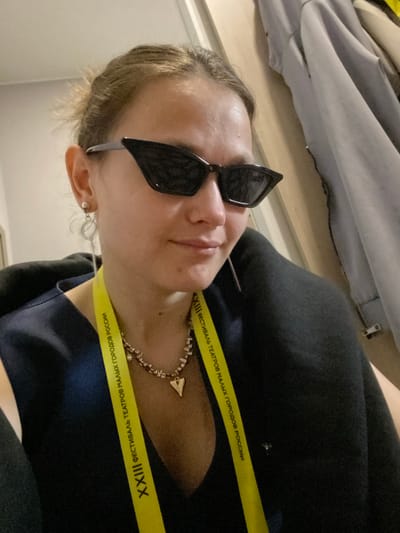 A selfie shows a woman wearing sunglasses and a yellow lanyard around her neck. The lanyard reads "XXIII Фестиваль театров малых городов России" which translates to "XXIII Festival of Small Towns Theatres of Russia". The background includes clothing and a wall. The city of Peresvet, Russia, is not directly visible in the image, but the lanyard indicates the location of the event.