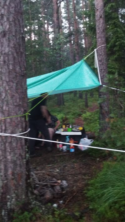A blurry, low-resolution image depicts a campsite in a forest. A turquoise tarp is strung between trees, providing shelter. Beneath the tarp, a makeshift table is laden with various items including containers, bottles, and a white plastic bag. Two individuals, one appearing to be wearing dark clothing and the other a patterned top, are visible behind the table, their faces obscured. The ground is covered with dirt, fallen leaves, and some debris, including what appears to be a small fire pit with embers. The surrounding environment is dense with trees and undergrowth. The image's quality and content do not provide any specific clues to indicate the location within the city of Tarasovskoe, Russia.