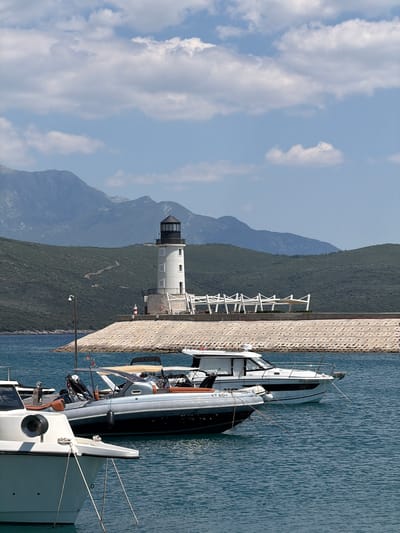 The image depicts a scenic view of a harbor in Radovići, Montenegro. The main subjects are a white lighthouse with a black cap and several boats docked alongside a stone pier. The lighthouse stands tall and distinct against a backdrop of lush green, mountainous terrain and a bright blue sky with scattered white clouds, suggesting a clear, sunny day.
The boats, varying in size and design, are moored to the pier, with their reflections subtly visible in the calm, blue water. One of the larger boats has "POLO00NIG" written on its side. Another boat has "KT 65H" visible. The pier itself is constructed of concrete blocks, providing a sturdy structure for the docking facilities. In the background, the rolling hills and mountains are densely covered with trees, adding to the natural beauty of the setting.
The scene appears to be set during the daytime, with ample sunlight illuminating the landscape and highlighting the details of the lighthouse and the boats. There are no people prominently visible in the image, focusing the attention on the architecture and the marine environment. The overall impression is one of tranquility and picturesque coastal scenery.