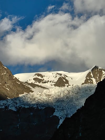 The image depicts a majestic mountain landscape. A vast, snow-covered peak dominates the center of the frame, with dark, rocky outcrops interspersed with white snow. Below the peak, a sprawling glacier is visible, characterized by its rugged texture and icy formations. The foreground is comprised of dark, shadowed mountains, contrasting sharply with the bright, snow-capped peaks. The sky above is a deep blue, with scattered white clouds adding depth and texture to the scene. The lighting suggests either early morning or late afternoon, casting dramatic shadows and highlighting the contours of the mountains and glacier. The overall impression is one of rugged natural beauty and imposing scale. There are no people or any signs of human activity in the image. The location is not specified as Kead, India.