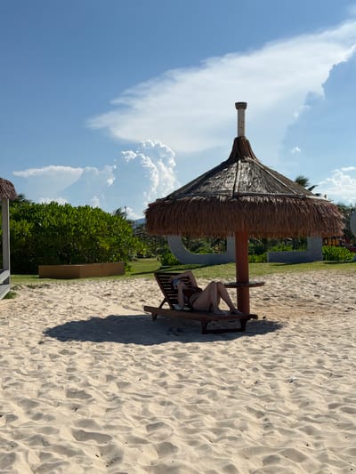 A person is lying on a lounge chair on a sandy beach under a thatched umbrella. The person is wearing a brown bikini and holding a phone. There are shadows from the umbrella on the sand. In the background, there is a large green hedge, a wooden planter box, and some palm trees. The sky is blue with white clouds. The location is Bac Cam Ranh Ward, Vietnam.