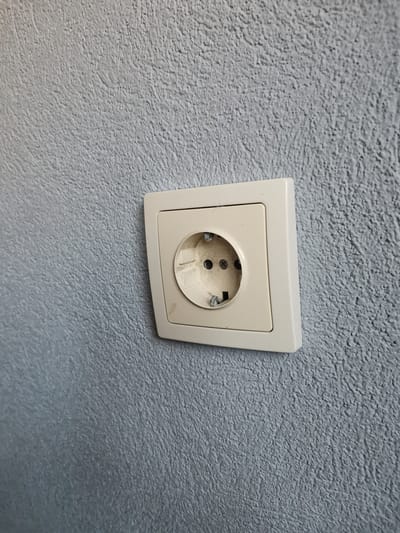 A single, beige-colored electrical wall socket, consistent with a Type F (Schuko) design, is mounted on a textured wall in Pokrovnik, Bulgaria. The wall displays an irregular, rough surface finish in a light gray or blue-gray hue. The socket's external plastic frame shows minor scuff marks and discoloration, particularly on the lower left side. The internal receptacle is visible, exhibiting wear and dirt around the two circular pin entry points and the metallic grounding clips positioned at the top and bottom. No electrical plug is inserted, and the socket is not actively in use. The environment is indoors, with diffuse, even lighting present. No individuals or other objects are depicted in the frame.