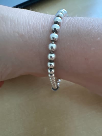 A close-up, angled shot shows a silver bead bracelet on a person's wrist. The bracelet is composed of evenly spaced, polished silver beads. The wrist is pale and shows visible skin texture. The background is a blurred, light brown surface. There is no information to suggest this image is located in the city of Murcia, Spain.