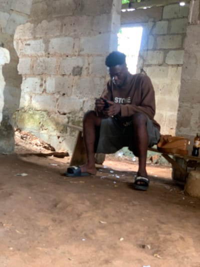 A young man wearing a brown long-sleeved shirt with the word "STONE" visible on it, grey shorts, and black slippers sits on a wooden bench, using his cell phone. His legs are bent at the knees, and his feet are flat on the floor. He is seated in a partly constructed building with concrete block walls and a dirt floor. The building appears to be under construction and has an opening in the wall. The man is looking down at his phone, and his right hand is holding it up to his ear. There is a brown box on the bench to the right of the man, and a bottle of what appears to be a dark liquid is sitting next to the box. There is some debris and dirt on the floor, and a small amount of vegetation is growing outside the building. The location is in Akure, Nigeria, with coordinates of latitude 7.225, longitude 5.175.