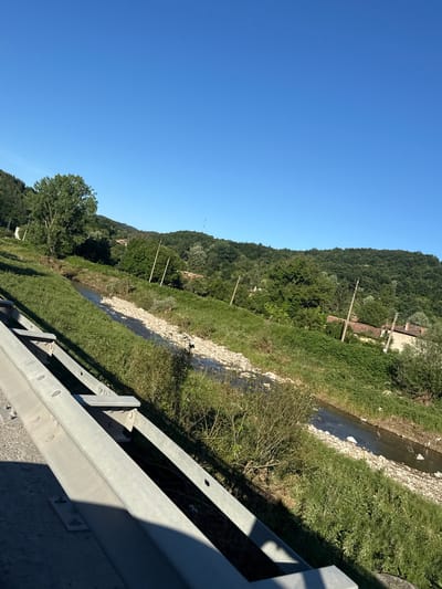 This media file displays a rural landscape in Veselinovo, Bulgaria, under a clear blue sky, suggesting daytime and fair weather. The scene is dominated by a lush, green hillside covered in dense trees. A shallow, rocky stream or riverbed winds its way down the slope, bordered by grass and low-lying vegetation. Several wooden utility poles with wires are visible along the hillside, indicating the presence of infrastructure in the area. In the mid-ground, a small, rustic building with a tiled roof peeks through the trees, hinting at rural habitation. In the foreground, a section of a metal guardrail runs along the right side of the frame, suggesting the viewpoint is from a road or elevated pathway. The guardrail is constructed of galvanized metal and casts a distinct shadow across the lower portion of the image. No people are visible in the scene. The overall impression is one of a peaceful, natural setting with some human elements integrated into the landscape.