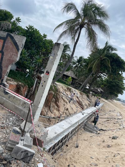 The image depicts a beach construction site in Phan Thiết, Vietnam, with a damaged concrete structure in the foreground. A large, partially collapsed concrete pillar stands to the left, adorned with graffiti. Red and white caution tape delineates the construction area, running across debris and the sandy ground. Several individuals are engaged in construction work, with one person in the middle ground pouring concrete into a mold, while others are nearby overseeing or assisting. Behind the construction area, palm trees and lush vegetation line the sandy shore, interspersed with thatched-roof structures that suggest a resort or beachfront establishment. The sky is overcast, indicating a cloudy day. The scene captures an ongoing effort to repair or reinforce the beachfront infrastructure amidst signs of erosion or damage.