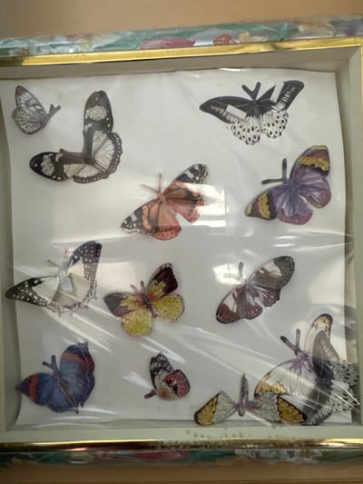 The image displays a framed collection of artificial butterflies. The butterflies are varied in color and pattern, with some appearing to be made of paper or fabric. They are arranged in a grid-like fashion within the frame, which has a gold border and is covered by a clear plastic sheet. The background within the frame is white. The overall presentation suggests a decorative item, possibly for display in a home or craft setting. The scene is indoors, with the lighting appearing to be ambient, casting soft shadows. The location is specified as Пролетарский, Russia, however, there are no visual cues within the image that allow for identification of a specific city, neighborhood, or landmark. The image is a close-up shot, focusing solely on the framed butterfly collection.