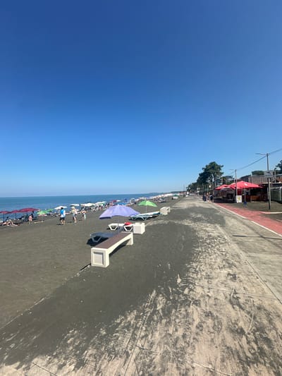 A clear, cloudless blue sky dominates the upper portion of the image. Below, a wide, dark sandy beach stretches along the Black Sea coast. Numerous beach umbrellas in various colors, including red, purple, green, and white, are set up along the shore, providing shade for beachgoers. Beach chairs and lounge benches are also visible.

Several people are scattered across the beach, some sitting under umbrellas, others walking or standing. The water of the Black Sea is calm and a deep blue.

To the right of the beach, there is a paved walkway or promenade. Along this walkway, there are several red-roofed kiosks or small shops, some with seating areas. Trees line the edge of the walkway further down, and buildings are visible in the distance. The walkway has a distinctive red stripe along its edge. The scene appears to be a lively beach resort in Ureki, Georgia, known for its magnetic sand beaches.
