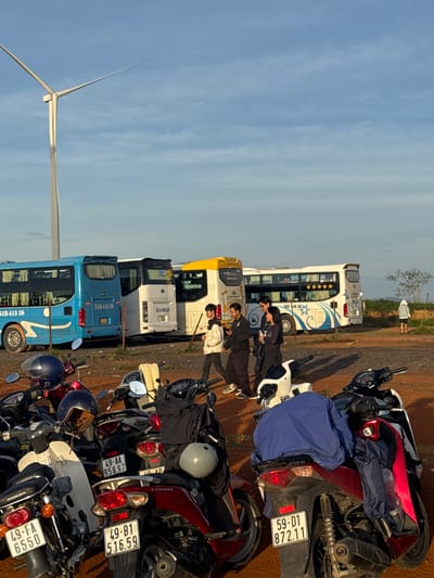 The image depicts a parking area with several motorbikes and buses, likely in Phát Chi, Vietnam, under a bright blue sky with sparse clouds. A prominent wind turbine is visible on the left side of the frame. In the background, three buses are parked, one blue, one white, and one yellow. A group of four individuals, two men and two women, are standing near the buses, seemingly engaged in conversation. One of the men is holding a stick. In the foreground, numerous motorbikes are parked, some with helmets and covers on them. The ground is covered in dirt and gravel. A lone person in a light-colored hoodie is visible in the far right background, standing near a field. The scene appears to be captured during daylight hours, possibly late afternoon due to the lighting.