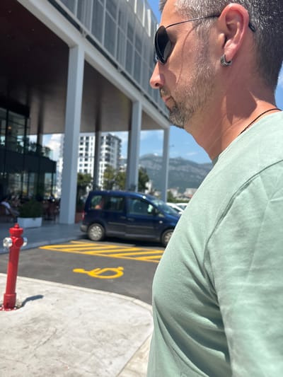 The media file shows a close-up of a man's profile, facing left. He has short, graying hair and a beard, and is wearing sunglasses with black lenses and a light green t-shirt. An earring is visible in his left ear. The background is blurred but suggests an outdoor urban setting. Behind him, a blue van is parked next to a yellow-lined parking spot marked with a wheelchair symbol. In the distance, buildings and hills are visible under a bright, sunny sky, suggesting daytime. A red fire hydrant is in the foreground. The scene appears to be in Bjeliši, Montenegro. No text is visible.