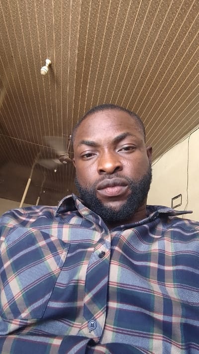 The image displays a close-up, low-angle portrait of an adult male of African descent, directly facing the camera with a neutral expression. He possesses short dark hair, a full dark beard, and a mustache. The subject is wearing a collared, long-sleeved plaid shirt with a pattern composed of dark blue, light blue, beige, and thin red lines. The top button of the shirt is open, and a buttoned chest pocket is visible on the left side.
The background depicts an indoor environment with a patterned ceiling composed of brown PVC panels, featuring a repetitive design of small star-like or geometric shapes. A small, off-white light fixture is mounted on the ceiling in the upper left portion of the frame. A three-bladed brown ceiling fan, blurred to indicate motion, is partially visible above and behind the subject's left shoulder. To the upper right, a plain, light-colored wall is visible, featuring a rectangular indentation or an empty frame. No specific visual identifiers within the indoor environment are present to confirm its precise location within the city of Muhammadu Dikko Stadium, Nigeria.