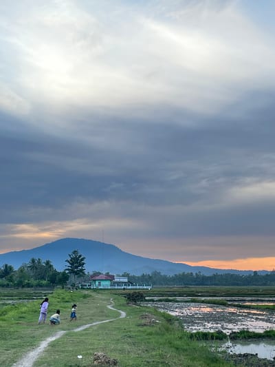 In Khang Tanjong, Indonesia, an outdoor scene unfolds under a cloudy sky with a bright, almost white patch of light near the top. A winding dirt path cuts through a grassy field where three young children are present. One child, wearing a light purple outfit, stands on the left. Another child, in a yellow and white outfit, is crouching down in the center. A third child, in a blue dress, is walking on the path to the right.

In the mid-ground, a cluster of trees, including a prominent tall green tree, stands beside a light green building with a red roof. Beyond the building and trees, a vast expanse of rice paddies stretches out, some containing water and mud, reflecting the warm hues of the sunset. A distant, misty blue mountain range forms the horizon, with a tall, thin structure, possibly a radio tower, visible near its peak. The overall environment appears rural and tranquil.