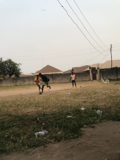 The image captures an outdoor scene in what appears to be a residential or semi-rural area, consistent with Umudike, Nigeria. The time of day is daytime, with an overcast or hazy sky providing soft, diffused lighting.
Three young males are visible, engaged in activity on an unpaved dirt path. In the midground, two boys are playing soccer; one boy, wearing a dark t-shirt and lighter shorts, is actively interacting with a soccer ball, while another boy in a yellow/orange t-shirt and dark shorts stands nearby, also seemingly part of the game. Further back, a third boy in a white and orange t-shirt and dark shorts stands near a building, observing or waiting.
The setting features a prominent dirt road or path, flanked by patches of green grass and scattered bits of litter, including plastic bags. In the background, simple, single-story buildings with sloped roofs (likely corrugated metal) are visible, partially enclosed by concrete block walls or fences. Overhead power lines and a utility pole extend across the upper left portion of the hazy sky, indicating basic infrastructure. There is no visible text in the image.