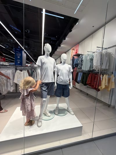 This is a medium shot of a clothing store display, taken from a high angle and reflected in glass. Two mannequins, one in a white t-shirt and denim shorts, and another wearing a similar outfit, are positioned on a white platform. A young blonde child in a patterned dress and tan Crocs stands between them, reaching out towards the lower mannequin. Behind the mannequins, clothing racks display various garments, including shirts and shorts in a range of colors. In the background, there's a poster with a woman's face and some signage, including what appears to be "FITTING ROOM," "KIDS," and "WOMEN." The lighting suggests an indoor environment, likely during the day. The overall scene depicts a retail space, with a child interacting with the display. Visible text includes "599.", "FITTING ROOM", "KIDS", "WOMEN", "VOVEECKT", "2208 MAVIO", and "SUNDAY". The location context is Novorossiysk, Russia.