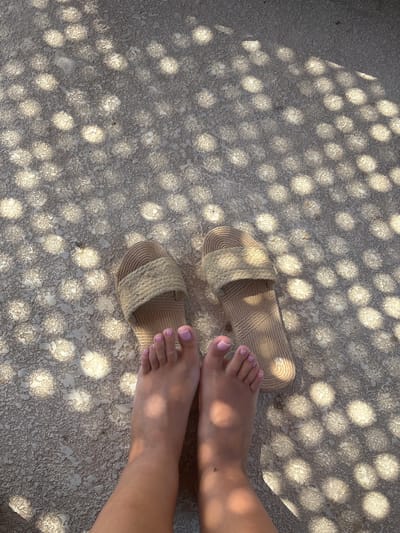 This media file captures a close-up, overhead view of a person's feet resting on a textured, light-colored surface. The surface is speckled with shadows that create a dappled light effect, suggesting the presence of sunlight filtering through an overhead structure, possibly a lattice or umbrella. Two light brown, woven sandals are placed on the surface, one to the left and slightly above the other, indicating they have been removed.
The subjects are the lower legs and feet of a person, with their toenails painted a soft pink color. The skin tone suggests a warm-weather setting. The scene is static, with no clear activity taking place beyond resting. The lighting and dappled shadows suggest it is daytime, likely sunny. The overall mood is relaxed and peaceful. There is no visible text in the image. The setting is indeterminate but the textures and lighting evoke a sense of being outdoors, perhaps in a patio, poolside, or a beachside area.
