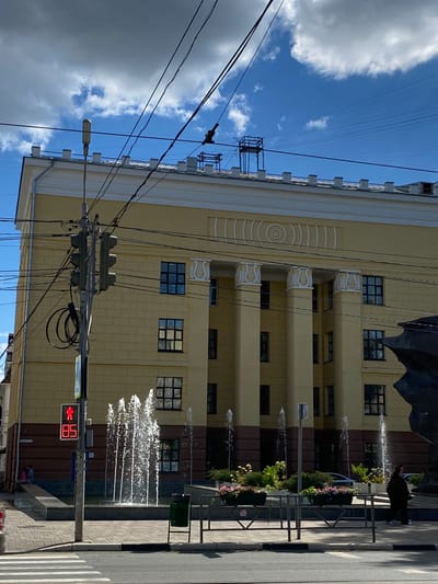 The image captures a street scene in Novokuybyshevsk, Russia. The primary subject is a tall, light yellow building with a patterned facade and multiple windows. In front of the building, a series of fountains are active, spraying water into the air.
A person, dressed in dark clothing, stands near the fountains, appearing to observe them. The scene is set outdoors on a sunny day, indicated by the bright light and the presence of fluffy white clouds against a blue sky. Power lines crisscross the upper portion of the frame, suggesting an urban or semi-urban environment.
Notable details include decorative elements on the building, such as moldings and a circular pattern on the left side. Flower planters are visible along the street, adding a touch of greenery and color. A pedestrian traffic signal displays a red "walk" symbol, indicating that pedestrians should not cross.
The overall mood of the image is one of a pleasant, ordinary day in a city. The fountains suggest a public space designed for leisure or aesthetic appeal. The time of day appears to be daytime, given the lighting conditions.