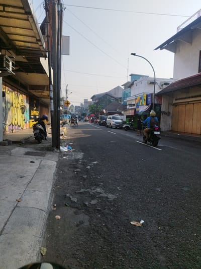 This is a street-level, medium shot of a road in Yogyakarta, Indonesia. The sun is low in the sky, casting long shadows. On the left side of the road, there is a sidewalk lined with buildings that feature graffiti on their lower sections. A dark-colored scooter with a blue helmet on its seat is parked on the sidewalk. The road itself is paved and appears to be wet in places. Several vehicles are on the road, including a white van, several motorcycles, and a three-wheeled vehicle. A person on a motorcycle is in the foreground, wearing a blue shirt and a backpack. There are also various shops and stalls visible along the roadside. The overall impression is of a busy urban environment.