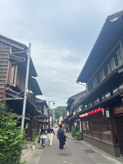 An overhead view captures a traditional street scene in Kanazawa, Japan, under an overcast sky. The image is oriented vertically, showing the sky at the top and bottom with buildings on either side, as if looking up a narrow street from a low angle. When viewed correctly, the perspective is down a historic street flanked by dark-stained wooden buildings with traditional tiled roofs and intricate latticework.
On the right side of the street, a row of red and white lanterns with floral patterns hangs from the eaves of a building, beneath a sign with Japanese characters and partially visible English text that might read "SAMURAI-GASHIRUYA-EN". Below the lanterns, two framed pictures are displayed on the wooden facade. The left side of the street features similar traditional wooden architecture, with prominent drainage pipes and gutters. A utility pole with a mounted camera is visible in the foreground on this side.
Several people are walking along the paved street, moving away from the viewer. One individual in a dark, possibly traditional, garment walks in the center. Behind them, two women in casual attire – one in a black top and white pants, the other in a black top, blue jeans, and a white ruffled undershirt, carrying a light brown bag – are visible. Green foliage, including bushes and potted plants, lines the sidewalk. In the distance, more buildings and a tree-covered hillside are visible under the muted light of the cloudy day. The overall atmosphere suggests a quiet, historic district.