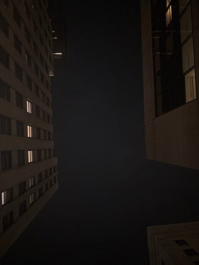 This image captures a low-angle, upward view of a dense urban environment at night in Boston, United States, likely within its central business districts such as the Financial District or Downtown Crossing.
On the left, a multi-story building with a light-colored, possibly beige or off-white, facade extends vertically out of the frame. This structure features numerous uniformly rectangular windows arranged in a grid pattern, with several windows on different floors displaying illuminated interiors with warm, yellowish-orange light. Above this building, a smaller, darker structure is subtly visible against the sky.
On the right, a second, distinct building dominates the frame, also extending vertically out of view. This structure has a darker, possibly grey or brown, facade with large, dark, reflective window panels. A few windows on this building also exhibit subtle interior illumination.
In the bottom right corner, a partial view of a third building is visible. This section features ornate architectural detailing on what appears to be a stone or paneled surface, suggesting an older construction style.
The expansive central and upper portions of the image consist of a uniform, dark, opaque sky, indicative of nighttime conditions. No celestial bodies or specific weather phenomena are discernible. There are no individuals, vehicles, or explicit actions depicted within the frame.