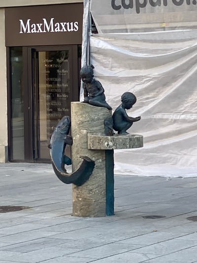 This image shows a bronze sculpture of two children and a fish, likely part of a fountain. The sculpture is positioned in front of a building with a storefront. One child is seated, appearing to interact with the fish, while the other child is leaning forward. The fish is depicted with its mouth open, curved upwards.
The building behind the sculpture has a large sign that reads "MaxMaxus" in white lettering on a dark brown background. The storefront windows reflect text that appears to be "MaxMara" and "SPORTMAX" repeated. This suggests the location is a fashion retail area.
A large, translucent white sheet is draped over scaffolding or construction materials behind the sculpture, obscuring the upper part of the building. The ground appears to be paved with light-colored stone tiles. The scene is likely set outdoors during the daytime, with natural light illuminating the area. The overall impression is of a public space with art and commercial activity.