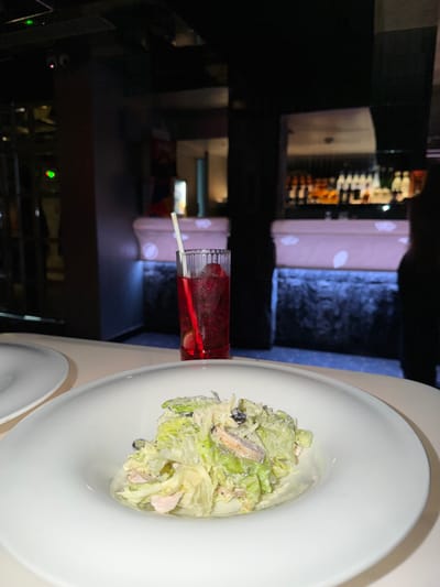 A high-angle, low-light photograph captures a dining setting within a modern establishment identified as New Delhi, India. In the immediate foreground, a white, wide-rimmed bowl contains a salad composed of chopped green lettuce, pieces of light pink meat (likely poultry or seafood), and a creamy dressing, topped with grated white matter. To the left, a portion of another white plate is visible. Behind the salad, a tall, clear, ribbed glass holds a red liquid, ice cubes, and what appear to be red fruit garnishes, with a white straw inserted. The table surface is light beige. The mid-ground and background are out of focus, revealing a dark environment. A bar counter with white panels featuring circular cutouts extends across the mid-background, illuminated by blue light strips at its base. Above the counter, shelves are stocked with numerous bottles, some reflecting light. On the far right, a dark, indistinct silhouette of a person is present. On the far left, blurred green and pink lights are visible. The overall scene depicts a prepared meal and drink in an ambient, dimly lit restaurant or bar setting.