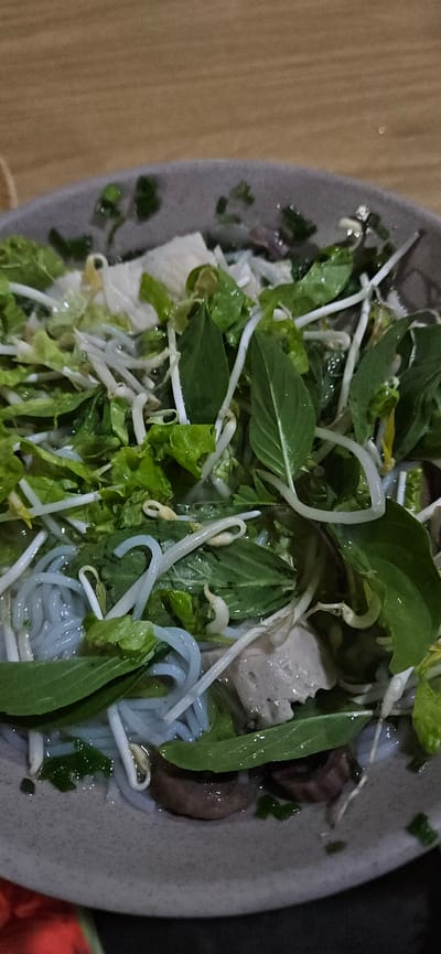 An overhead close-up shot captures a bowl of Vietnamese noodle soup or salad, likely a regional specialty from Cam Đức, Vietnam. The bowl, which appears grey or light brown, is filled with a vibrant mix of ingredients. Prominently featured are light blue or greyish thin noodles, suggesting a unique variety like Bún Đậu Mắm Tôm or a similar rice vermicelli dish.
A generous topping of fresh green herbs, including what looks like basil leaves and lettuce, along with numerous white bean sprouts, covers the surface of the dish. Beneath these greens, various protein components are visible: light-colored, somewhat irregular chunks that could be tofu or a type of processed meat, and darker, brownish pieces which might be mushrooms or a different meat. Chopped green onions or chives are also scattered throughout.
The bowl rests on a light brown wooden surface, possibly a table, which is visible on the left side of the frame. The lighting is soft and even, suggesting an indoor setting. No specific time of day or weather can be inferred. No people or text are visible in the image. The composition focuses tightly on the bowl, highlighting the textures and freshness of the ingredients.
